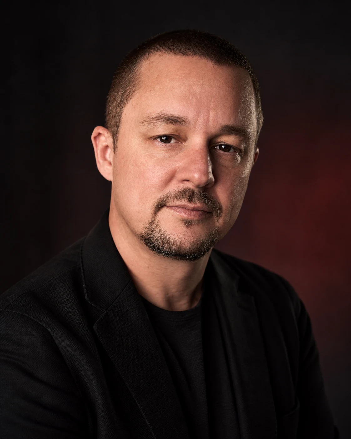 Portrait of Tony Simons, host of Less Lip, More Ship, in a dark jacket against a dark red background